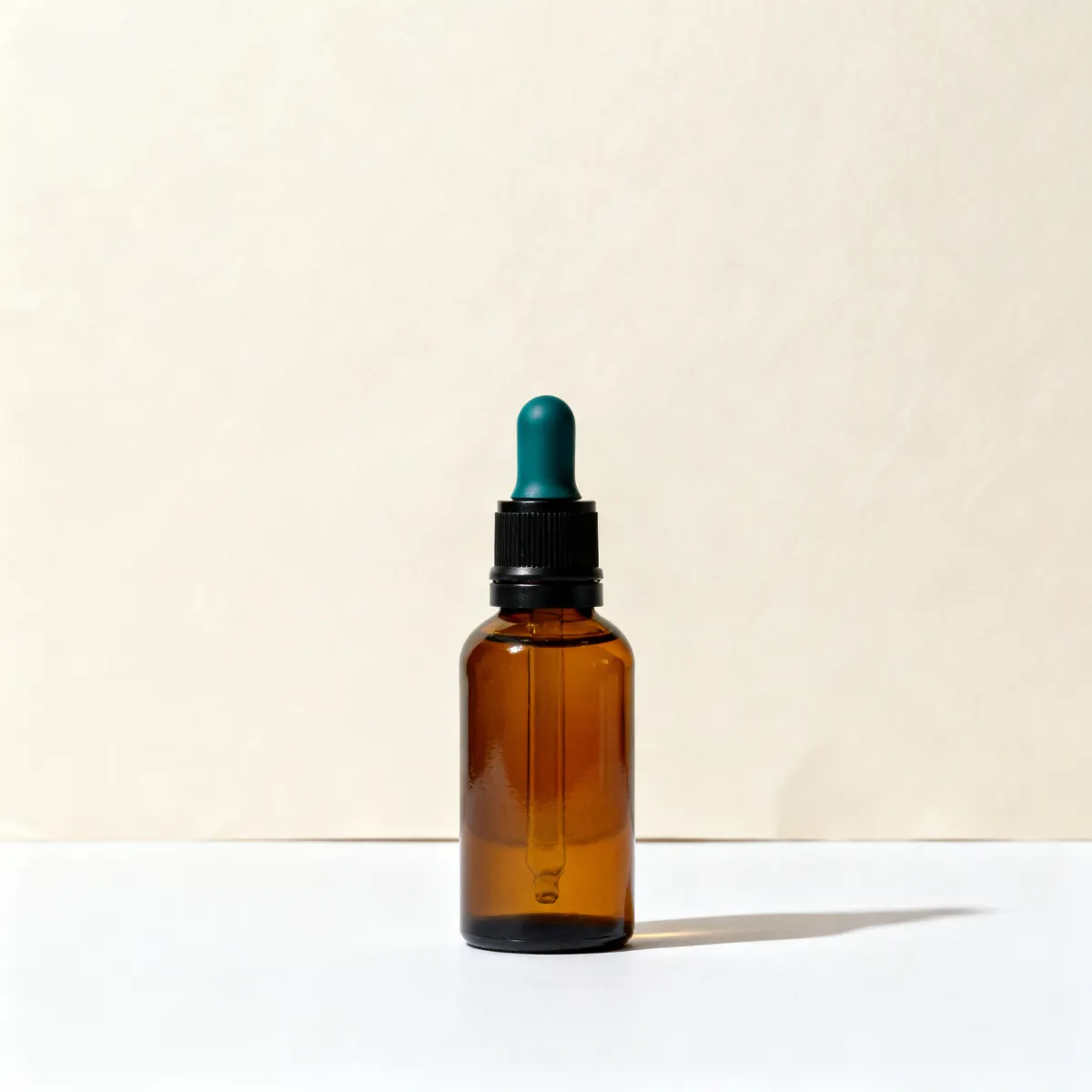 An amber glass dropper bottle with a teal bulb, centred on a warm-white seamless cove