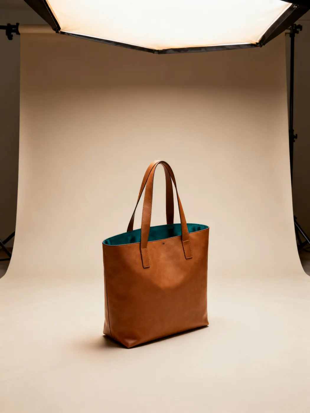 Tan leather tote standing upright, a teal lining just showing, softbox lighting the cove.