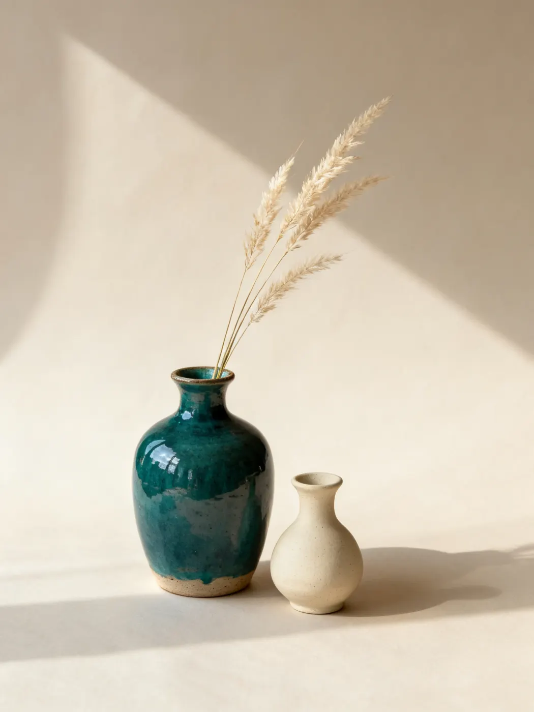 Teal-glazed stoneware vase with dried grass, a small cream bud vase beside it.